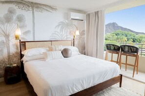 A bedroom with a large bed, two bedside lamps, a view of mountains, and a balcony with chairs. - Ocean Breeze Bungalow Across Waikiki Beach (Honolulu)