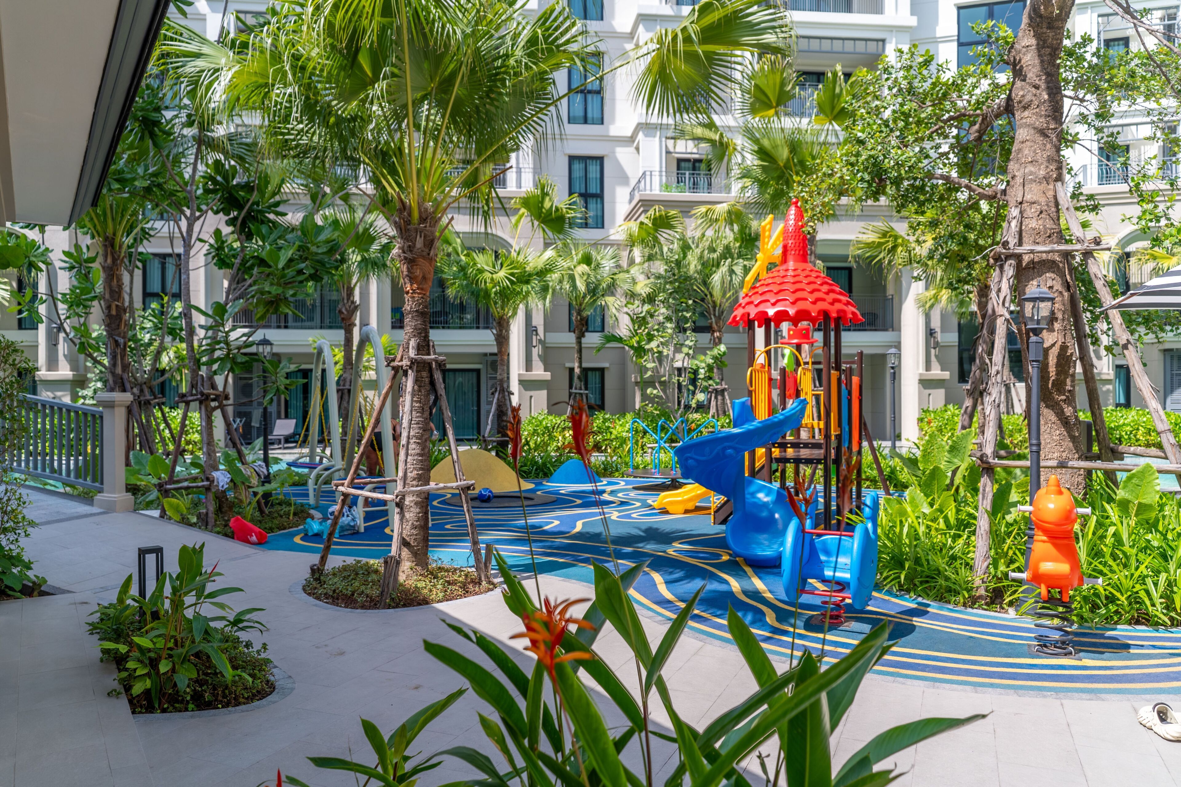 Uma área de recreação infantil com escorregadores e estruturas de escalada, cercada por palmeiras e um edifício ao fundo.