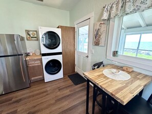 A compact kitchen with a stainless steel refrigerator, a washing machine, and a wooden dining table with a placemat. - Two Waterfront Cottages in Cedar Key Amazing View! (Cedar Key)