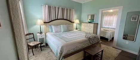 Quarto com cama, mesas de cabeceira, uma cadeira, ventoinha no teto e uma janela.