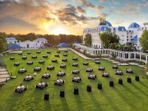 A large outdoor area with multiple round tables arranged on a lawn, surrounded by white buildings with blue roofs. - Taj Gandhinagar Resort and Spa (Mansa)