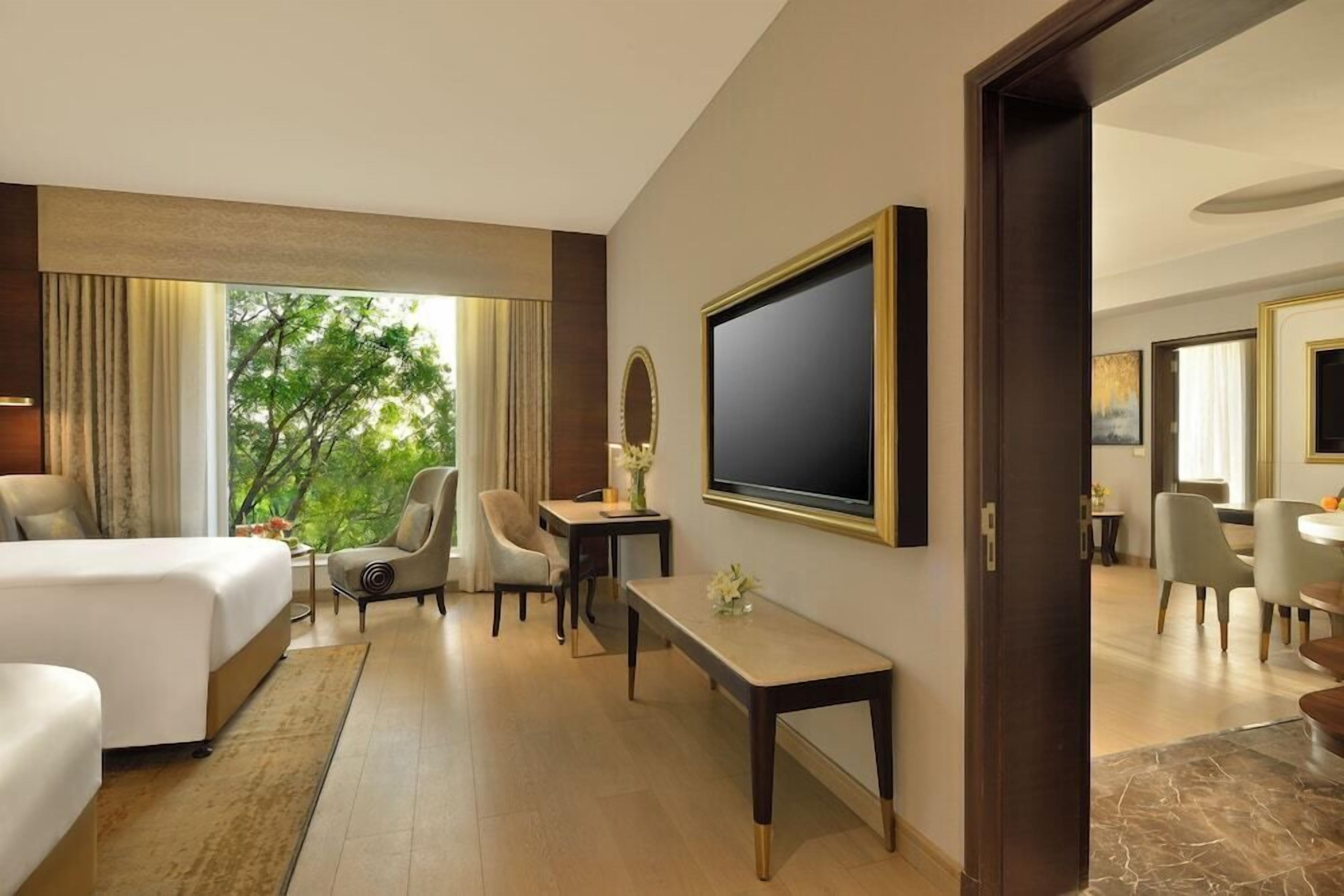 A hotel room with a bed, a desk, a chair, a TV, and a view of trees through the window.