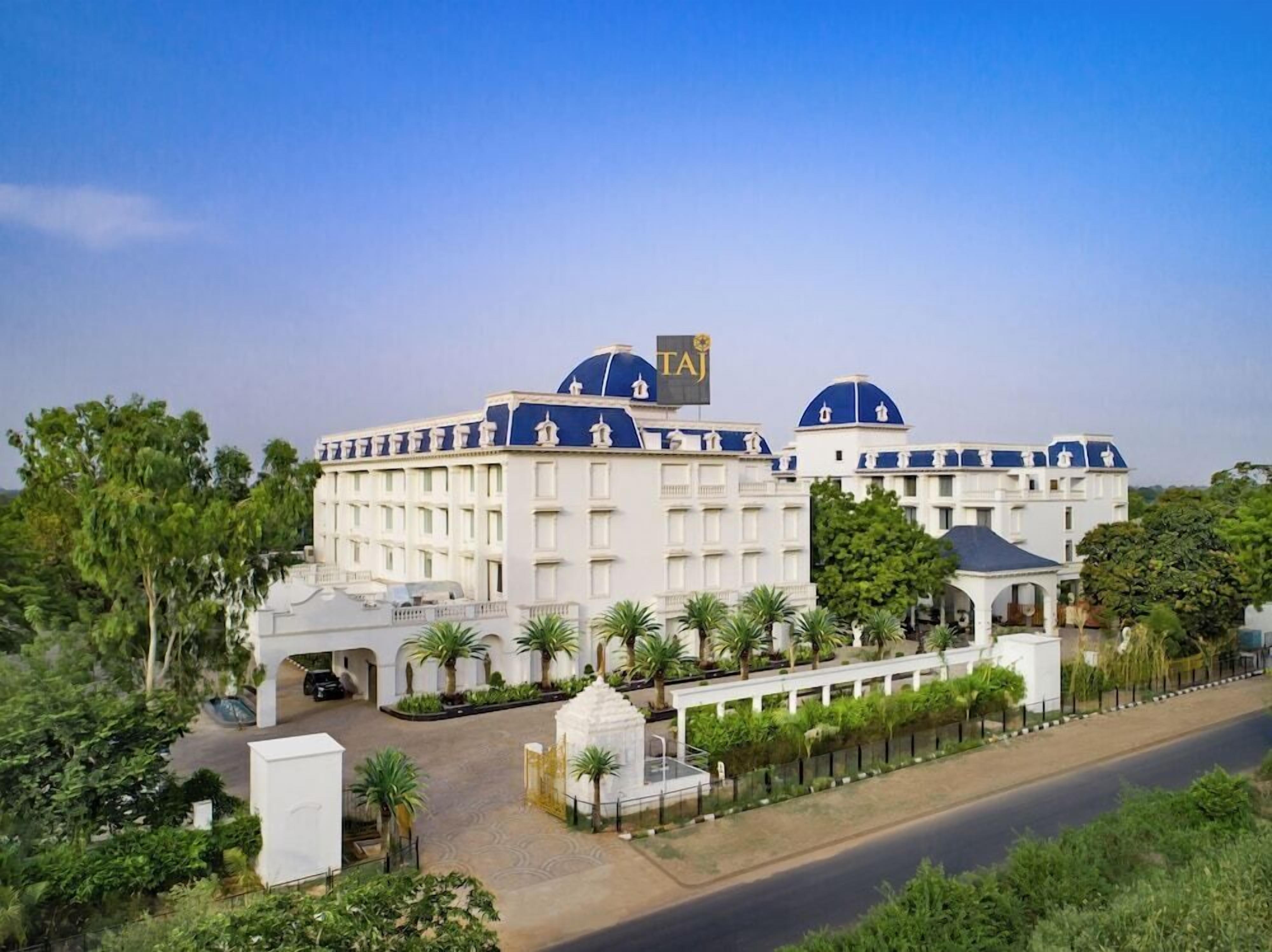 A large hotel with multiple white buildings and blue domes, a flag with 'TAJ' on top, and a well-maintained garden area.