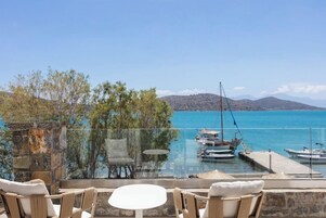 A terrace with a glass balustrade overlooking a marina with boats, a clear sky, and distant mountains.