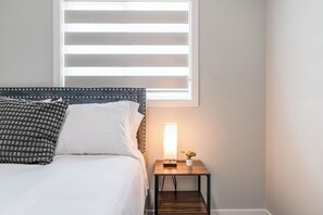A bed with a headboard, a bedside table with a lamp, and a window with blinds. - Stylish 3BR Home With A/C | Fast Wifi | Garage Parking | Sleeps 10 (Calgary)