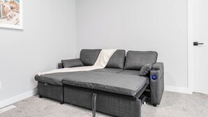 Een grijze bank met een witte plaid en twee kussens, geplaatst tegen een witte muur met een lijstwerkfoto.