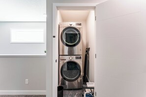 A laundry room with two stacked front-loading washing machines, a drying rack, and a small shelf with items. - Stylish 3BR Home With A/C | Fast Wifi | Garage Parking | Sleeps 10 (Calgary)