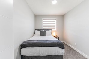 A bedroom with a bed, bedside table, lamp, and window with blinds. - Stylish 3BR Home With A/C | Fast Wifi | Garage Parking | Sleeps 10 (Calgary)