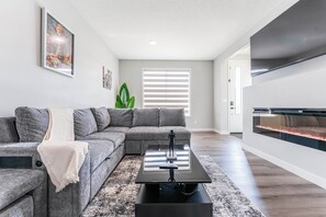 A modern living room with a sectional sofa, a coffee table, a fireplace, and a large TV. - Stylish 3BR Home With A/C | Fast Wifi | Garage Parking | Sleeps 10 (Calgary)