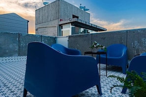 Eine Dachterrasse mit blauen Stühlen, einem kleinen Tisch mit einer Pflanze und einem Betongebäude im Hintergrund.