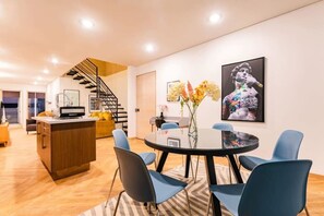 Ein modernes Interieur mit Essbereich, der einen runden Tisch und blaue Stühle umfasst, einer Küche mit einer Holzinsele und einer Treppe, die zu einer oberen Ebene führt.