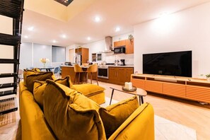Ein modernes Wohnzimmer mit einer Couch, einem Essbereich mit Stühlen, einer Küche mit Schränken und Geräten sowie einem Fernseher an der Wand.