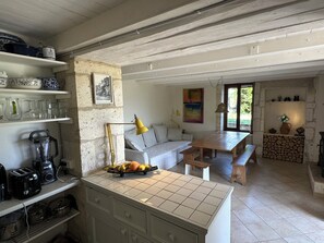 A kitchen with a wooden table, a bench, and a fruit bowl. There is a white cabinet with drawers and a shelf with glassware. A yellow lamp hangs from the ceiling. - Traditional 19th century stone farmhouse sympathetically restored with views. (Saint-Martial-Viveyrol)