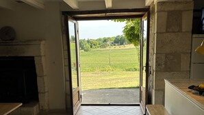 A room with a tiled floor, a fireplace, a kitchen area, and an open door leading to a view of a green field. - Traditional 19th century stone farmhouse sympathetically restored with views. (Saint-Martial-Viveyrol)