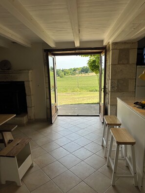 A room with a tiled floor, a fireplace, a kitchen area, and an open door leading to a view of a green field. - Traditional 19th century stone farmhouse sympathetically restored with views. (Saint-Martial-Viveyrol)