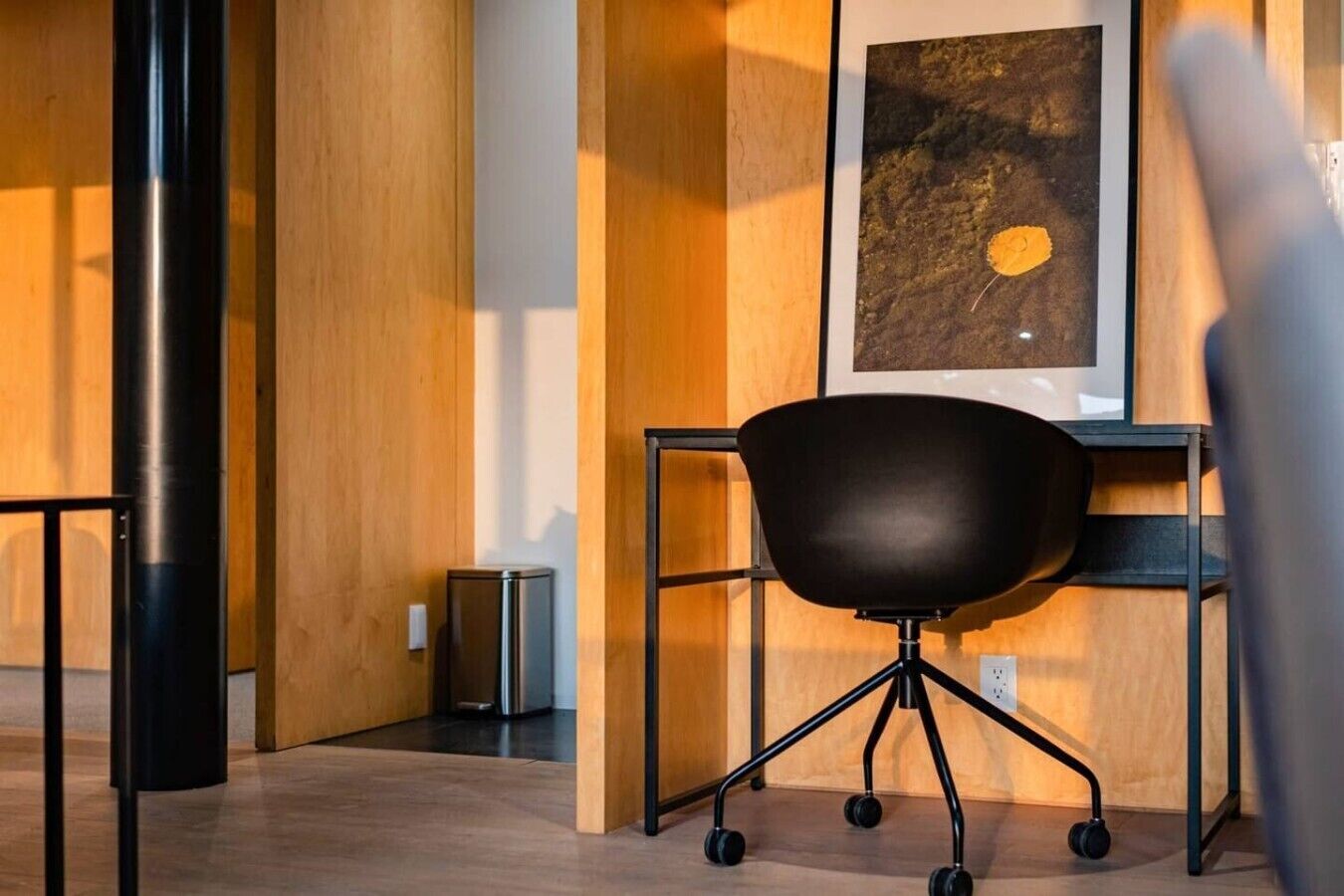 Un espace de bureau moderne avec un fauteuil de bureau noir, un bureau en métal et une œuvre d’art abstraite encadrée accrochée au mur.