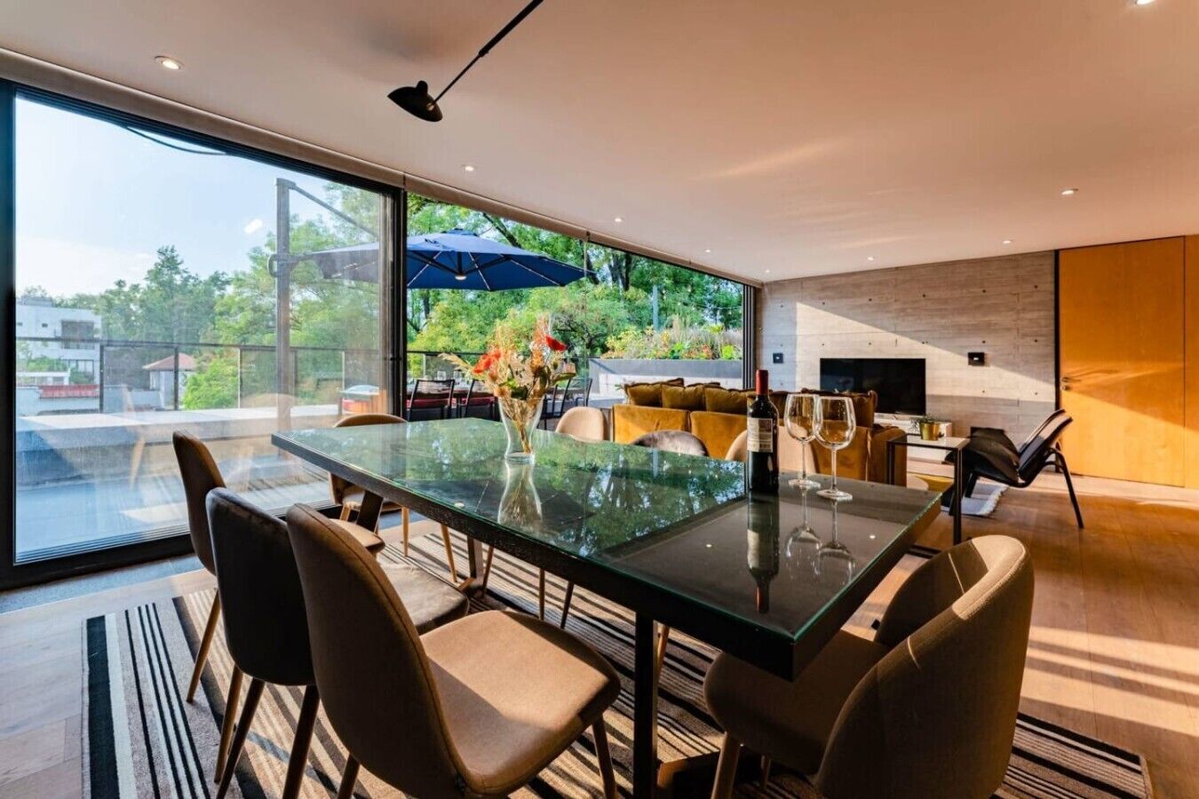A modern dining area with a glass table, chairs, and a view of greenery outside.