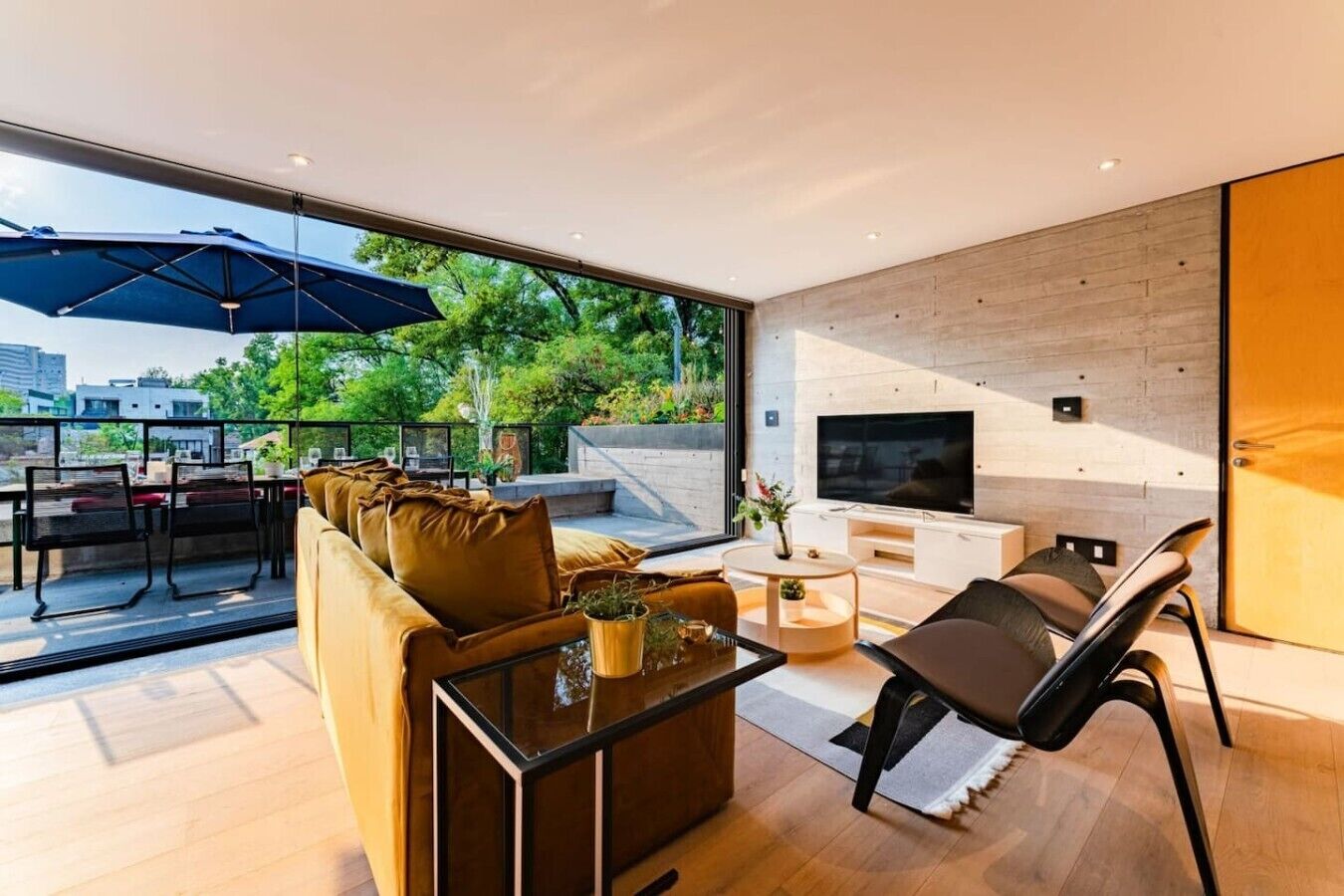 A modern living room with a sofa, coffee table, and TV. There is a balcony with outdoor furniture and a large umbrella.