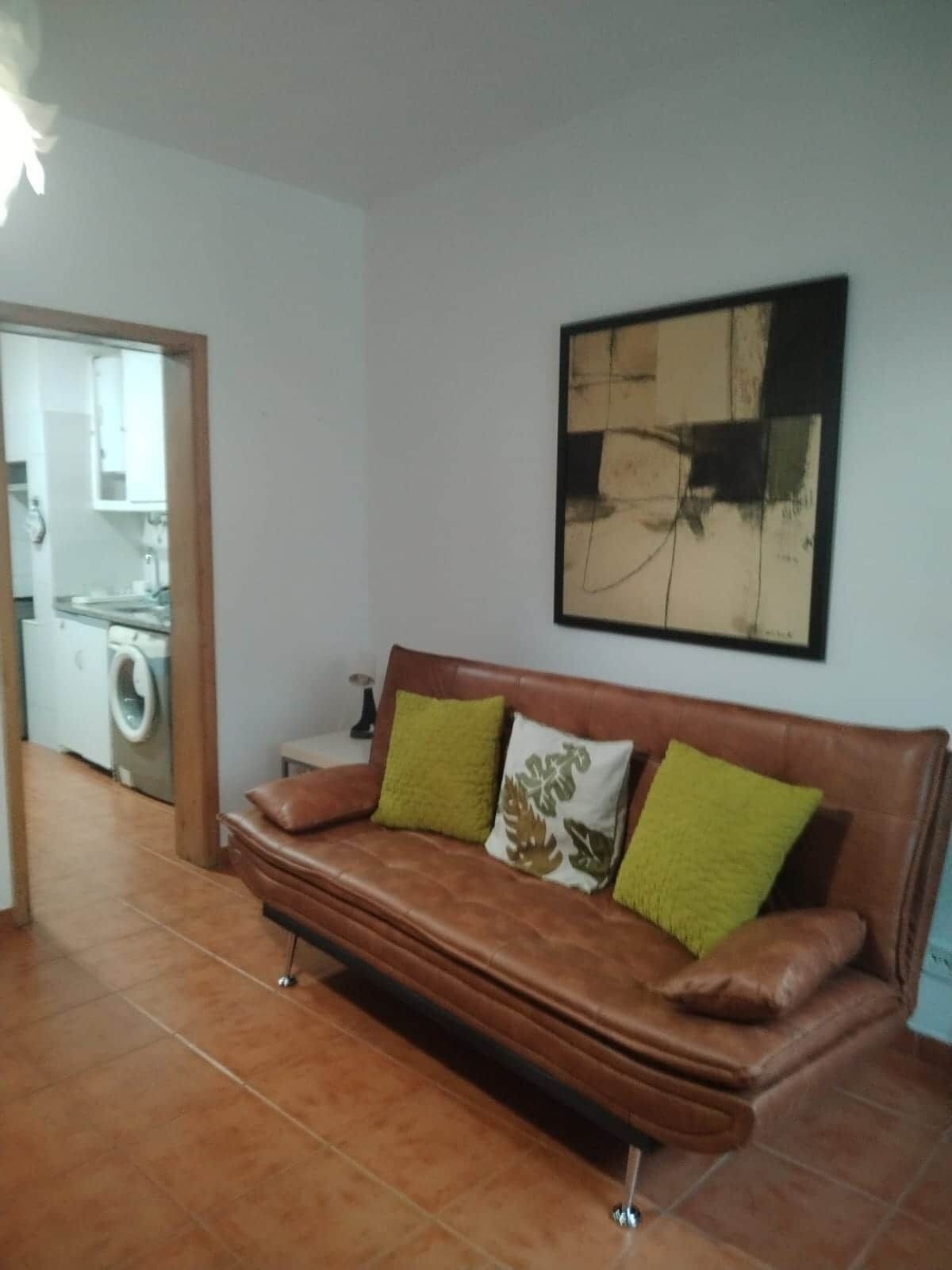 Una sala de estar con un sofá de cuero marrón, cojines verdes y estampados, piso de baldosas y un cuadro en la pared.
