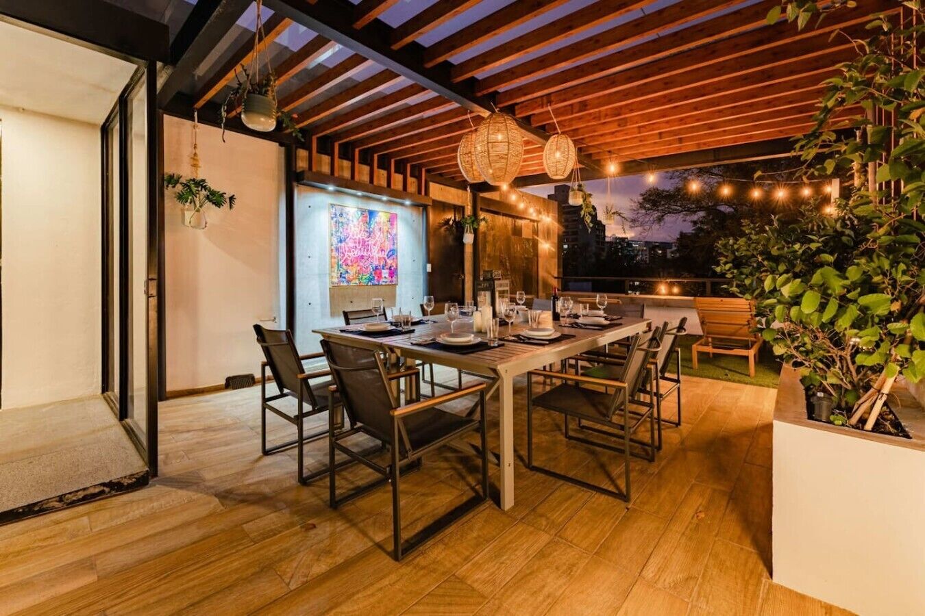 An outdoor dining area with a wooden table set for six, surrounded by chairs, under a wooden pergola with hanging lights.