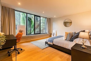 A modern bedroom with a large window, a bed with multiple pillows, a desk with a chair, and a small table with a lamp.