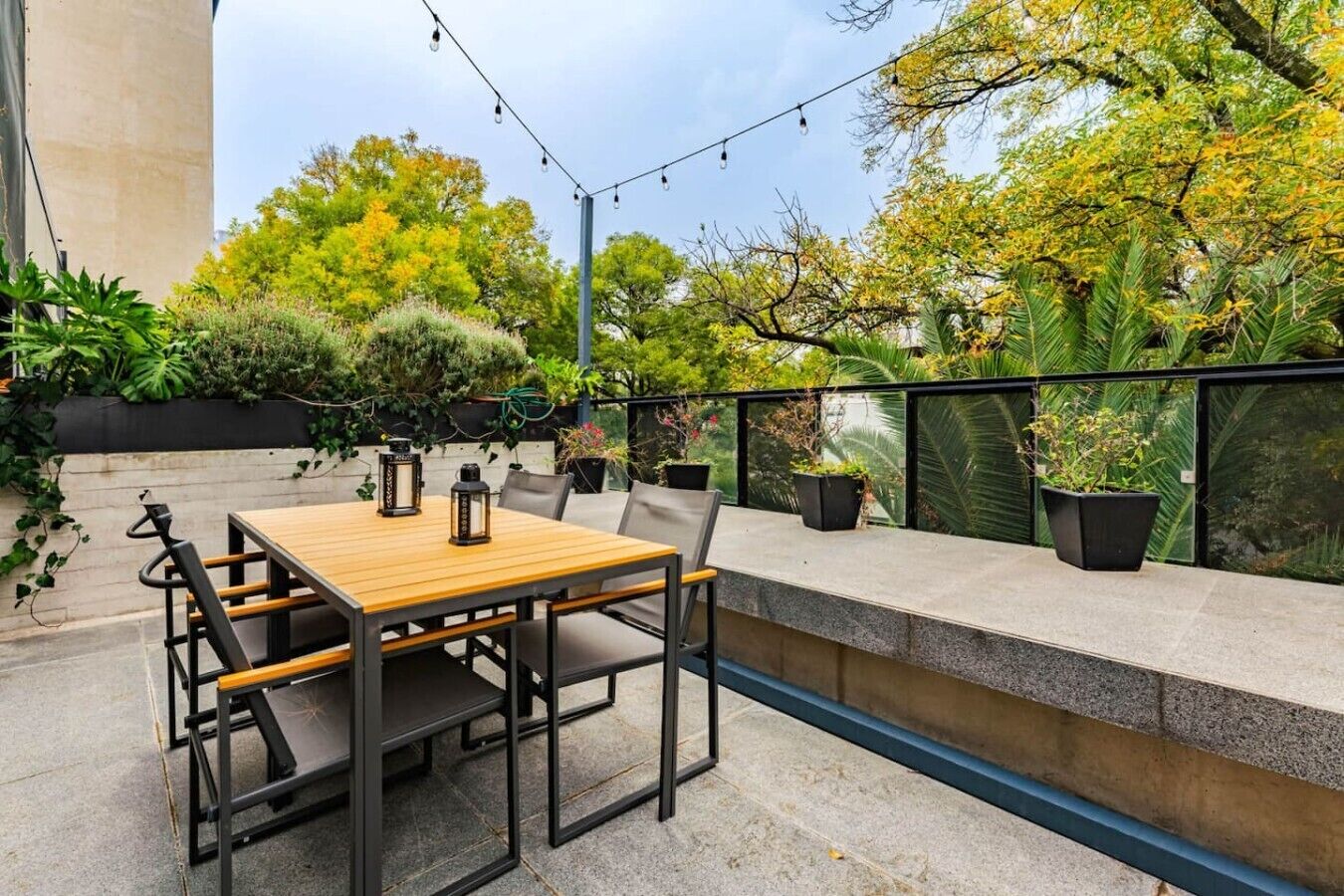 Une terrasse extérieure avec une table et des chaises en bois, entourée de plantes en pot et d’une rambarde.