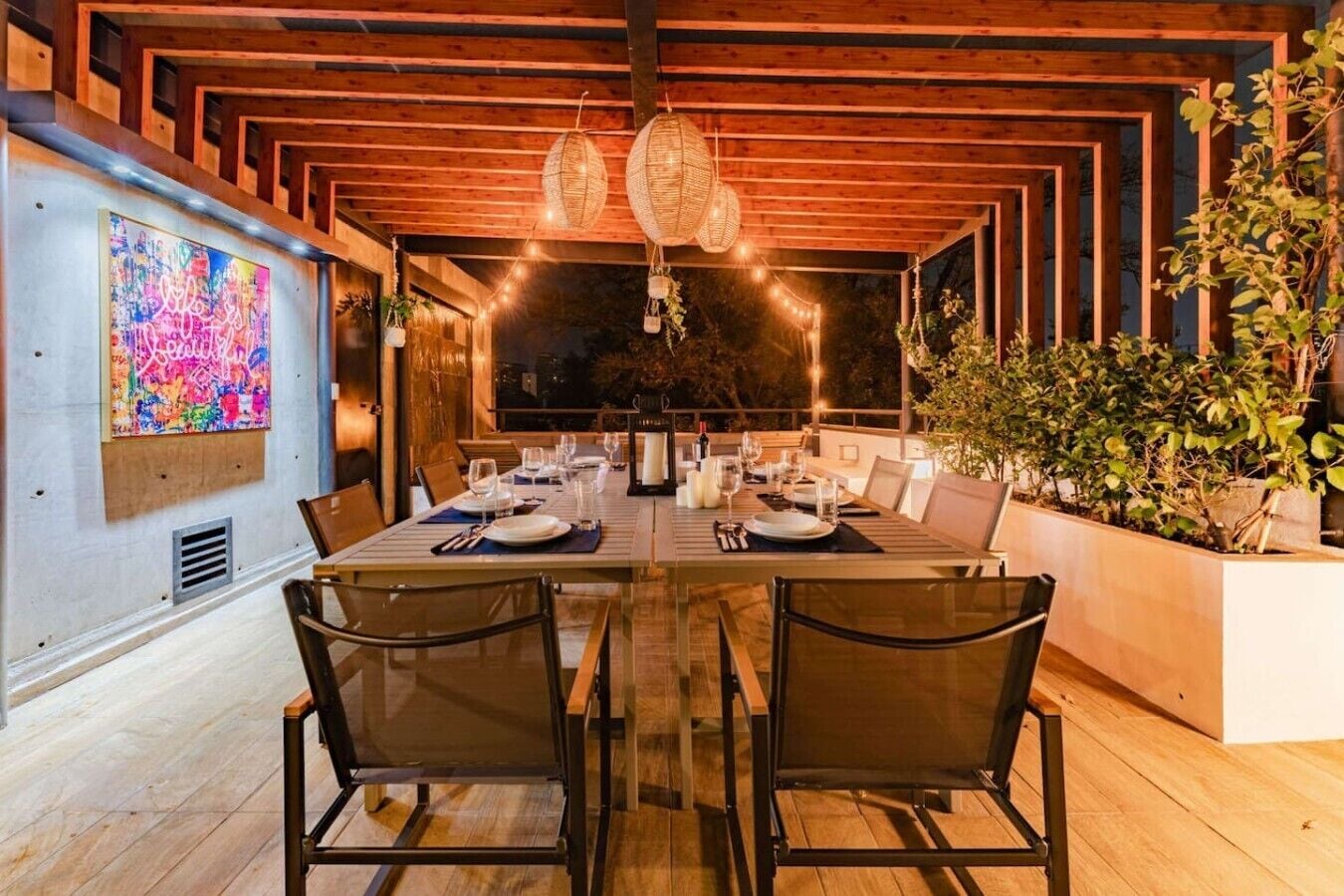 A dining area with a long table set for multiple people, wooden ceiling beams, hanging light fixtures, and a large abstract painting on the wall.