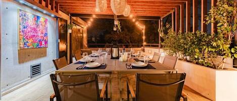 A dining area with a long table set for multiple people, wooden ceiling beams, hanging light fixtures, and a large abstract painting on the wall.