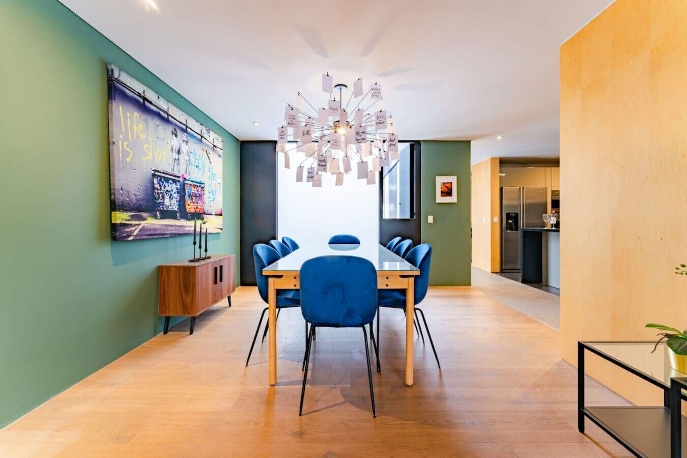 Un comedor moderno con una mesa de madera y sillas azules, una lámpara decorativa en el techo y un mural colorido en la pared.