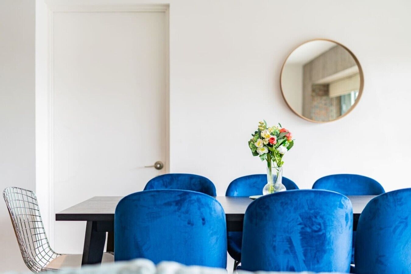 A dining area with a white door, a round mirror on the wall, and a table with blue chairs.