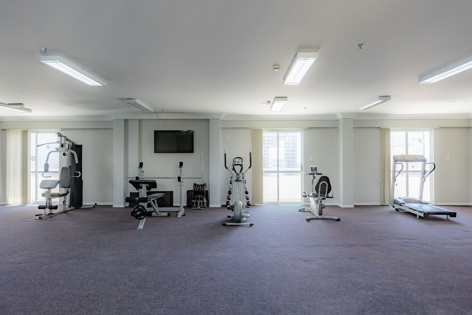A spacious gym with various exercise equipment, large windows, and a flat-screen TV on the wall.