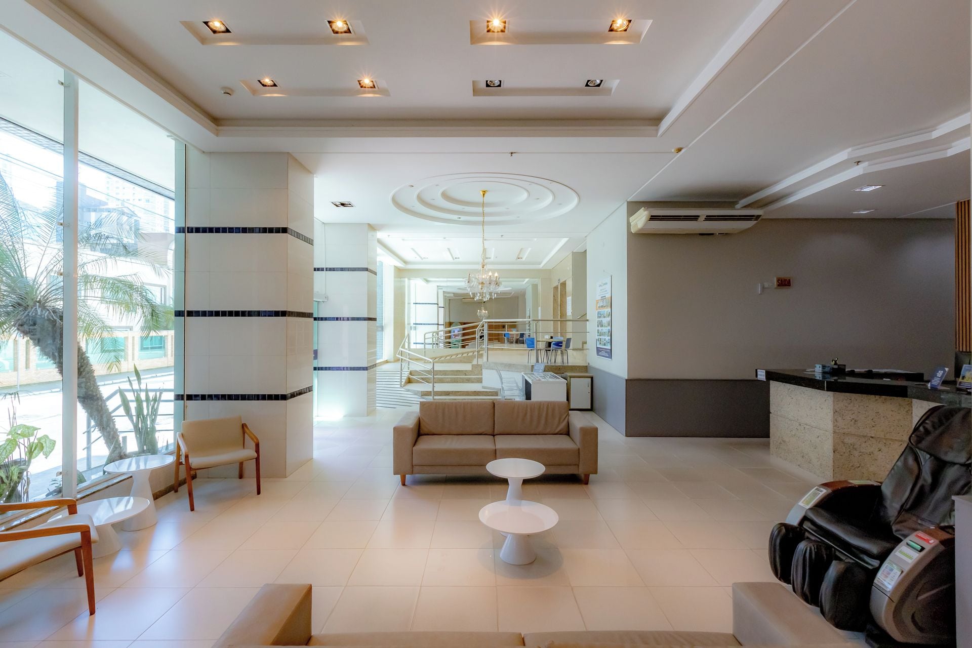 Um hall de hotel com balcão de recepção, área de estar, poltronas de massagem e escadas que levam aos andares superiores.