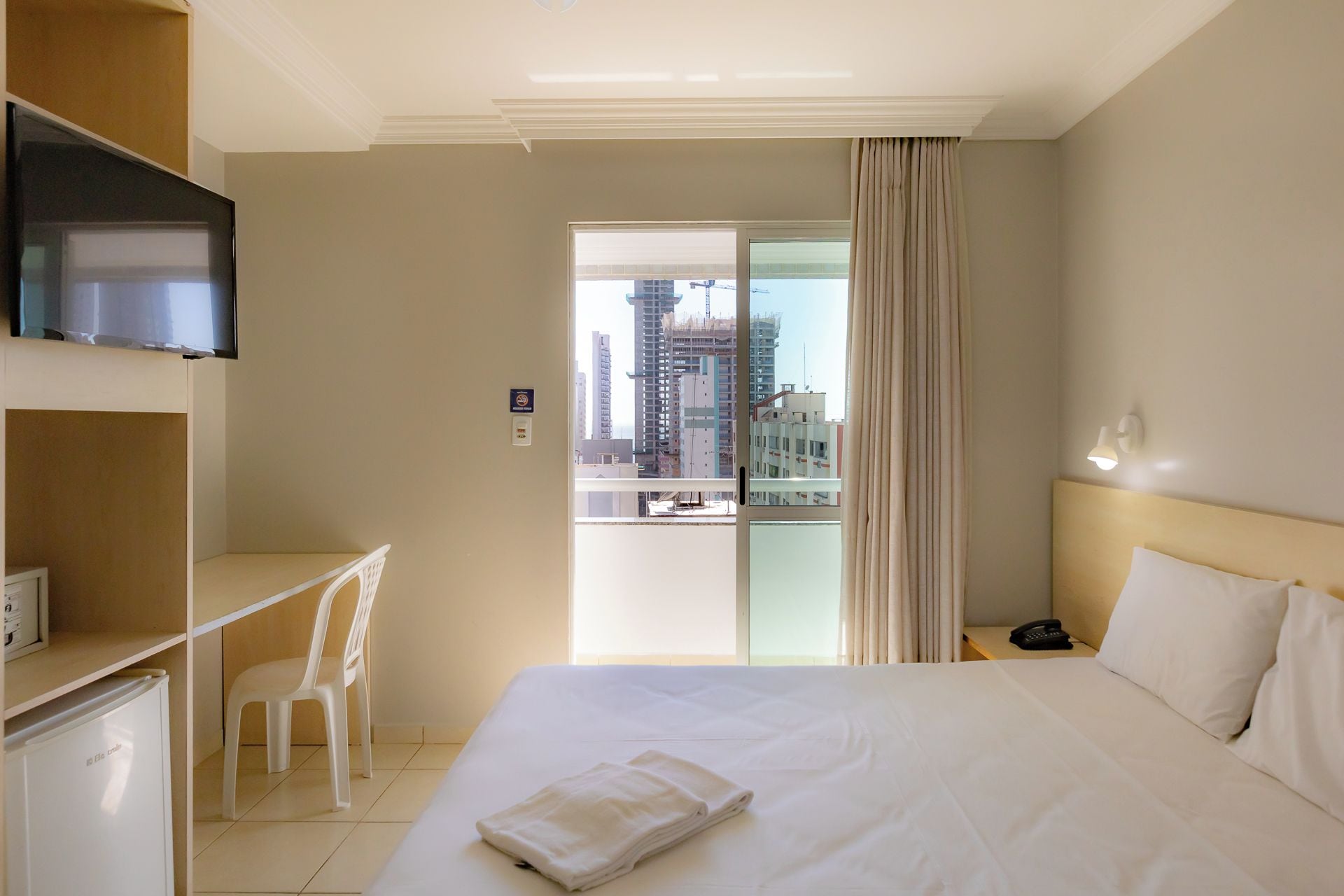 Habitación de hotel con cama, escritorio, silla, televisor, balcón y vista a la ciudad, además de un teléfono en la mesita de noche.