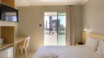 Habitación de hotel con cama, escritorio, silla, televisor, balcón y vista a la ciudad, además de un teléfono en la mesita de noche.