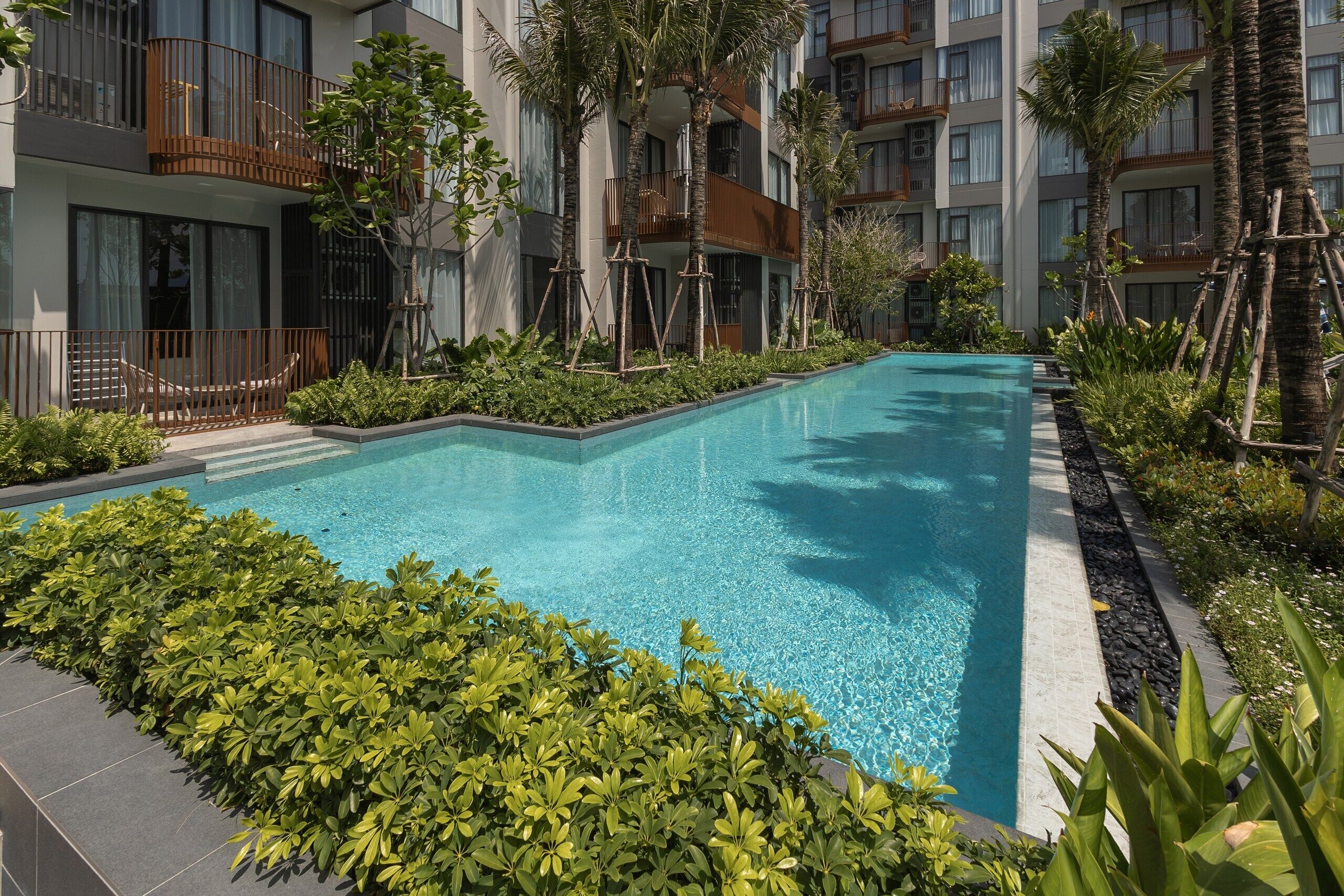 A rectangular swimming pool surrounded by lush greenery and modern buildings with balconies.
