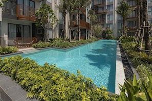 A rectangular swimming pool surrounded by lush greenery and modern buildings with balconies. - New 2BR Apartment at Sansiri Canvas, Bang Tao (Choeng Thale)
