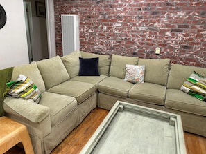 A L-shaped sectional sofa with multiple cushions, a wooden floor, and a brick wall. - Santa Monica two bedroom apartment SM5 (Santa Monica)