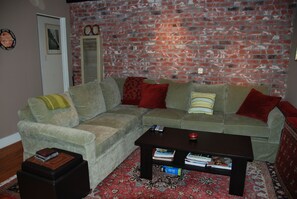 Ein Wohnzimmer mit einem Ecksofa, einem Couchtisch mit Büchern, einem gemusterten Teppich und einer Ziegelwand.