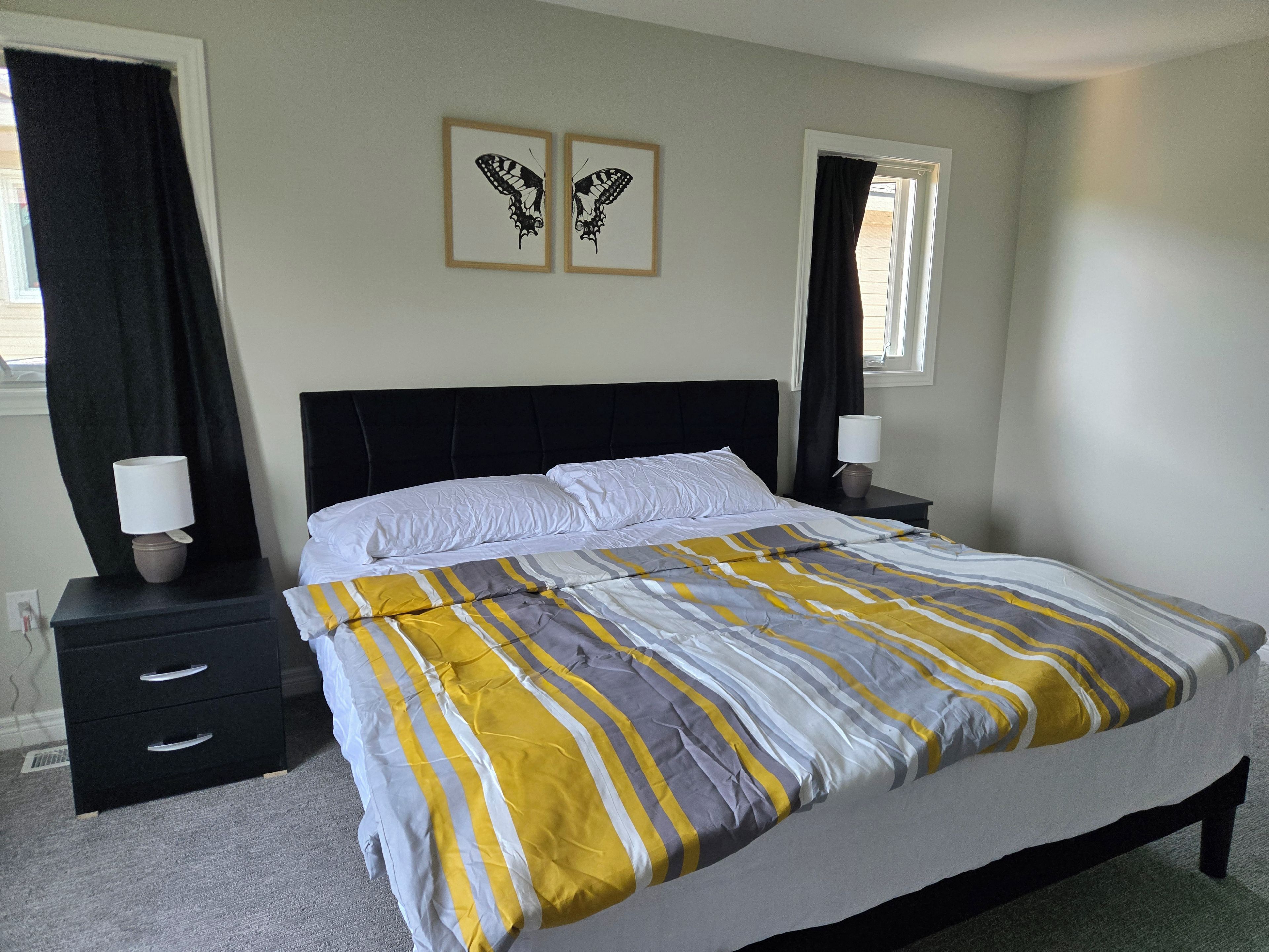 Een net opgemaakt bed met een gestreept laken, twee nachtkastjes met lampen, en twee ingelijste vlinder afbeeldingen aan de muur.
