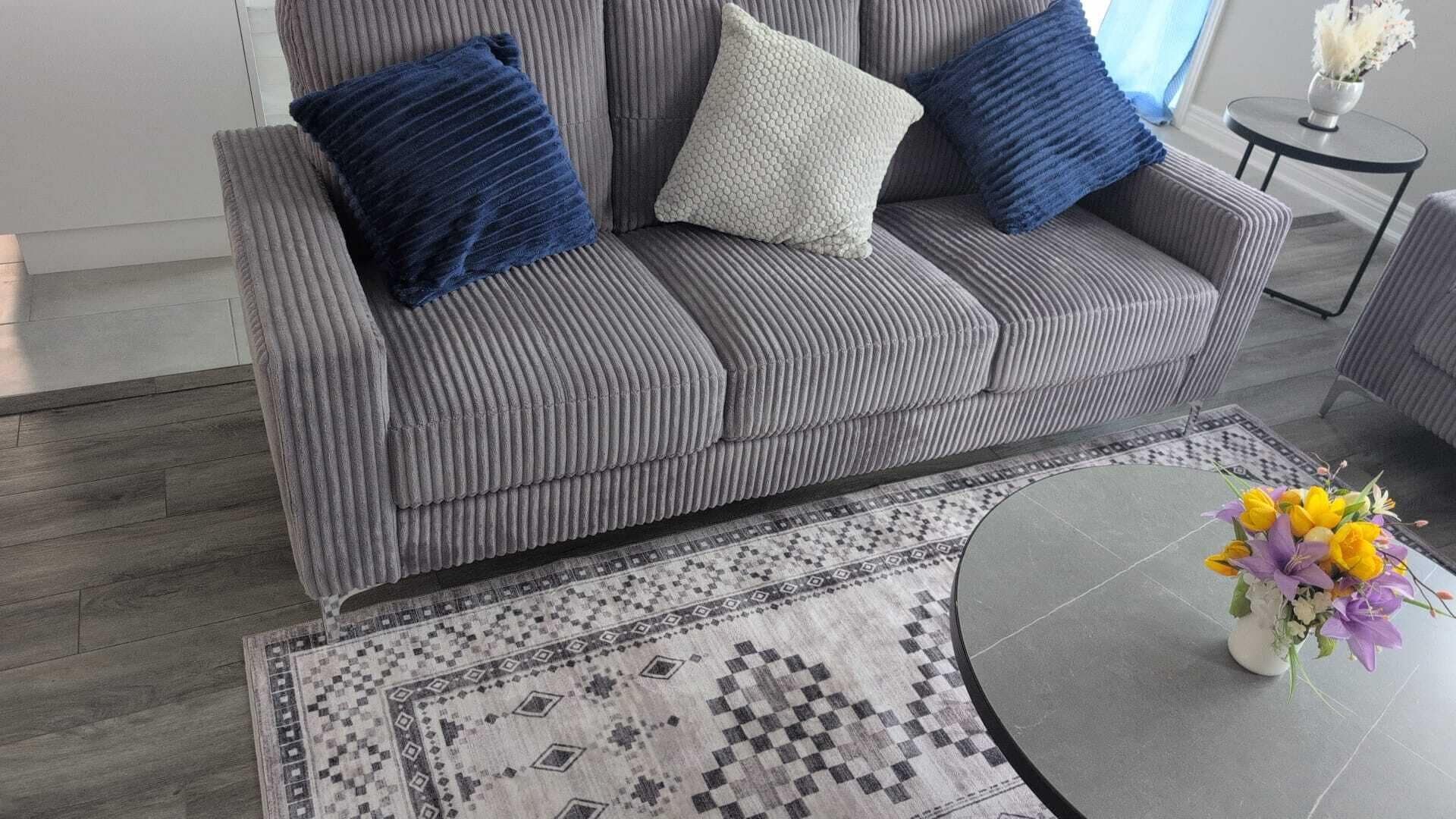 Een grijze gestoffeerde bank met een kamerkleed en een klein, rond tafeltje met een bloemenvaas.