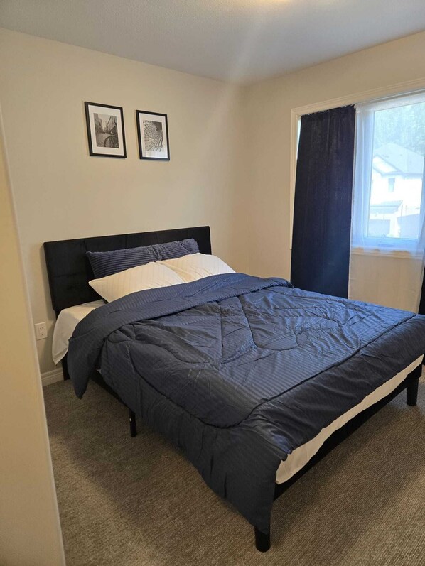 A single bed with a dark headboard and a quilted bedspread, two framed pictures on the wall, and a window with a dark curtain.