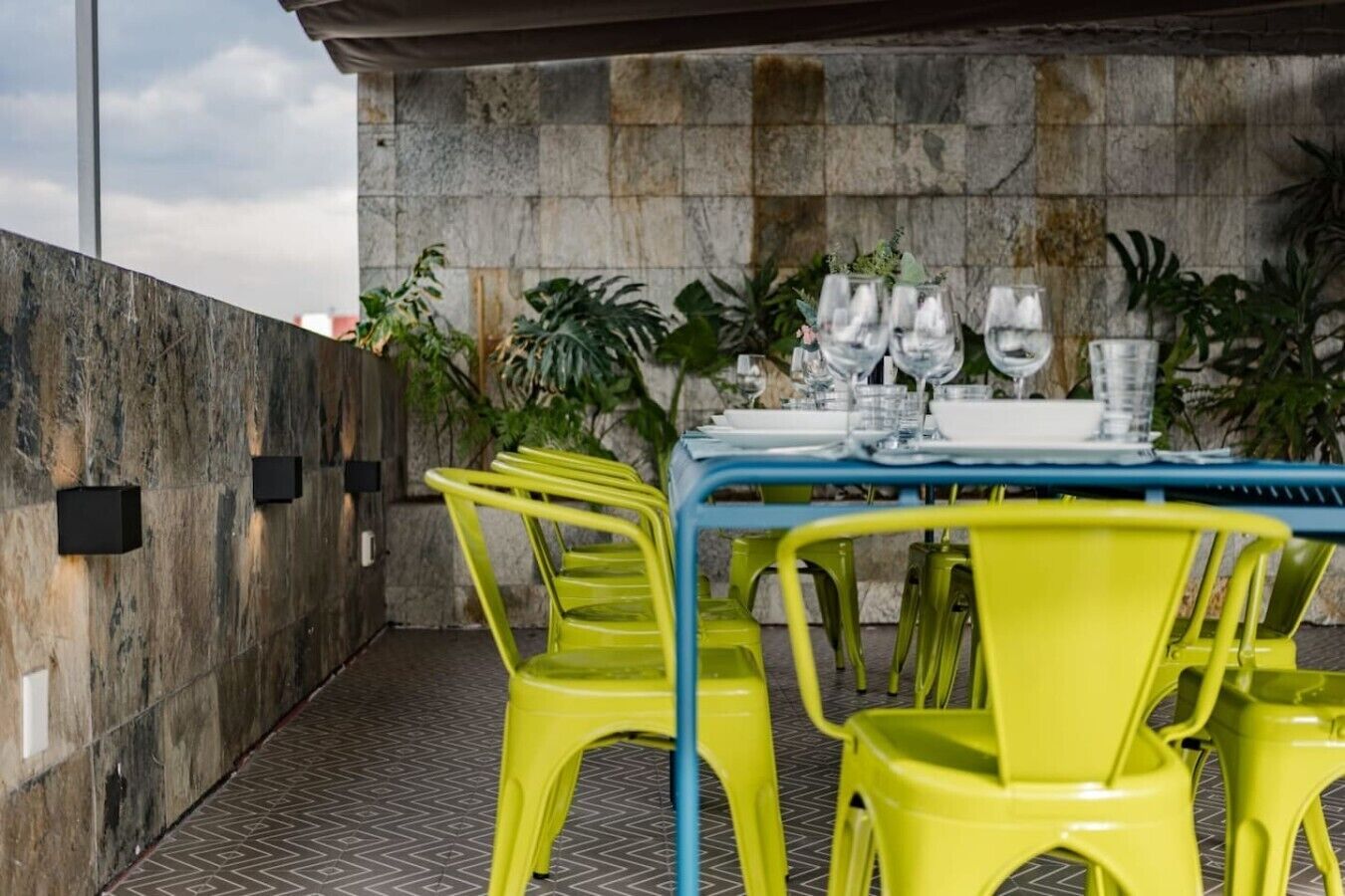 Area pranzo all'aperto con tavolo imbandito per quattro persone, circondato da sedie gialle e con una parete in pietra sullo sfondo.