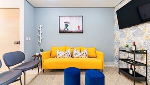 A living room with a sofa, two blue ottomans, a TV, and a shelf with glasses and a bottle. - 2 Condos in La Roma | Parking, Perfect for Groups (Mexico City)