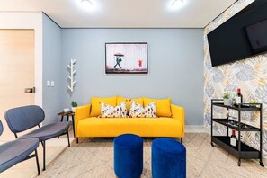 Ein Wohnzimmer mit einer Couch, zwei blauen Hocker, einem Fernseher und einem Regal mit Gläsern und einer Flasche.