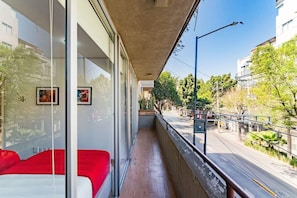 A balcony with a red sofa, glass walls, and a view of a street with trees and buildings. - 2 Condos in La Roma | Parking, Perfect for Groups (Mexico City)