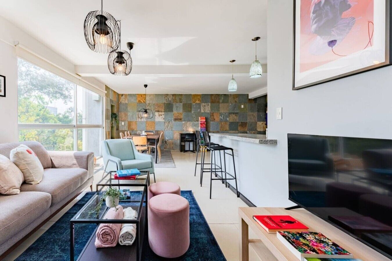 A modern living room with a sofa, coffee table, and TV, a dining area with chairs, and a kitchen with a stone backsplash.