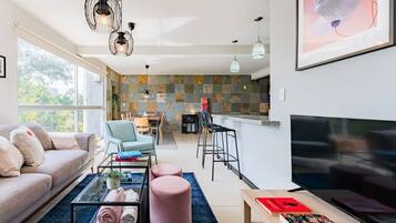 A modern living room with a sofa, coffee table, and TV, a dining area with chairs, and a kitchen with a stone backsplash.