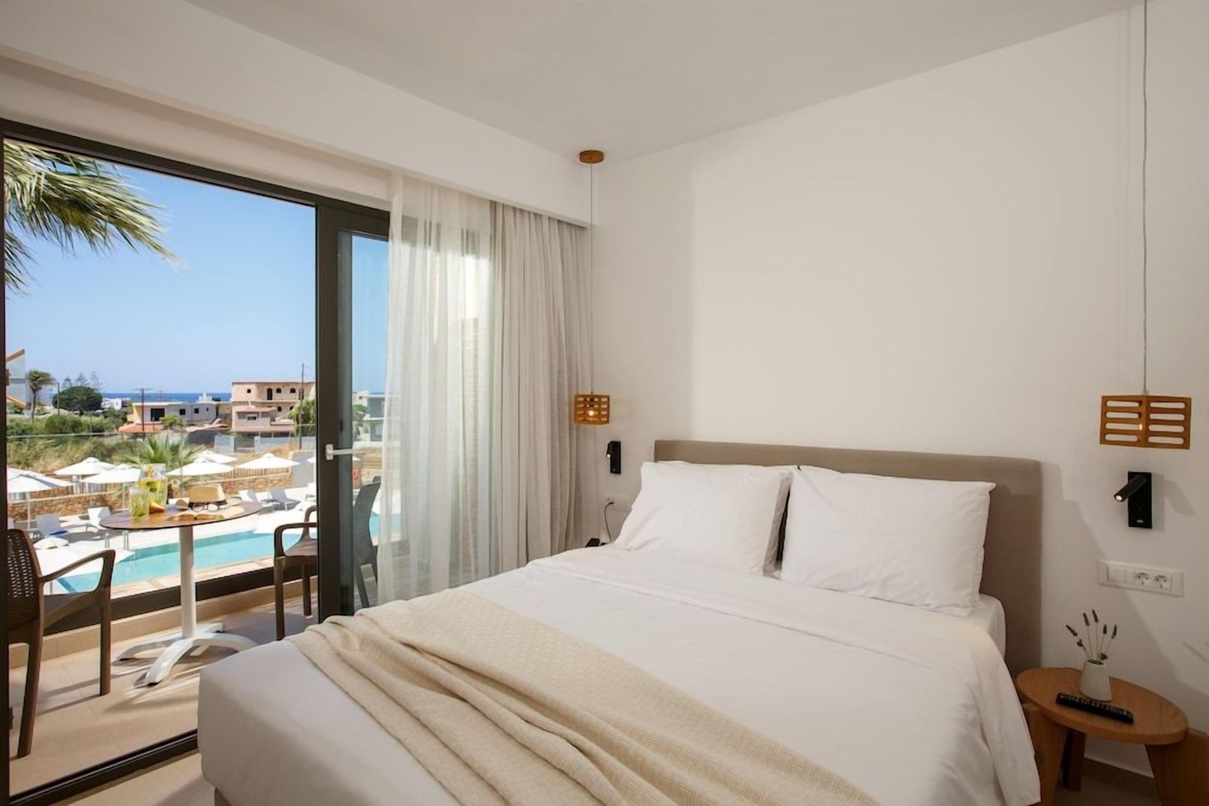 A hotel room with a large bed, a view of a pool and buildings, and a balcony with chairs.