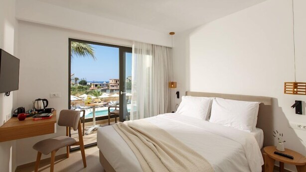 A hotel room with a bed, a desk with a chair, a TV, and a view of a pool and buildings. - Aloe Boutique Hotel Powered by Anissa Beach (Hersonissos)