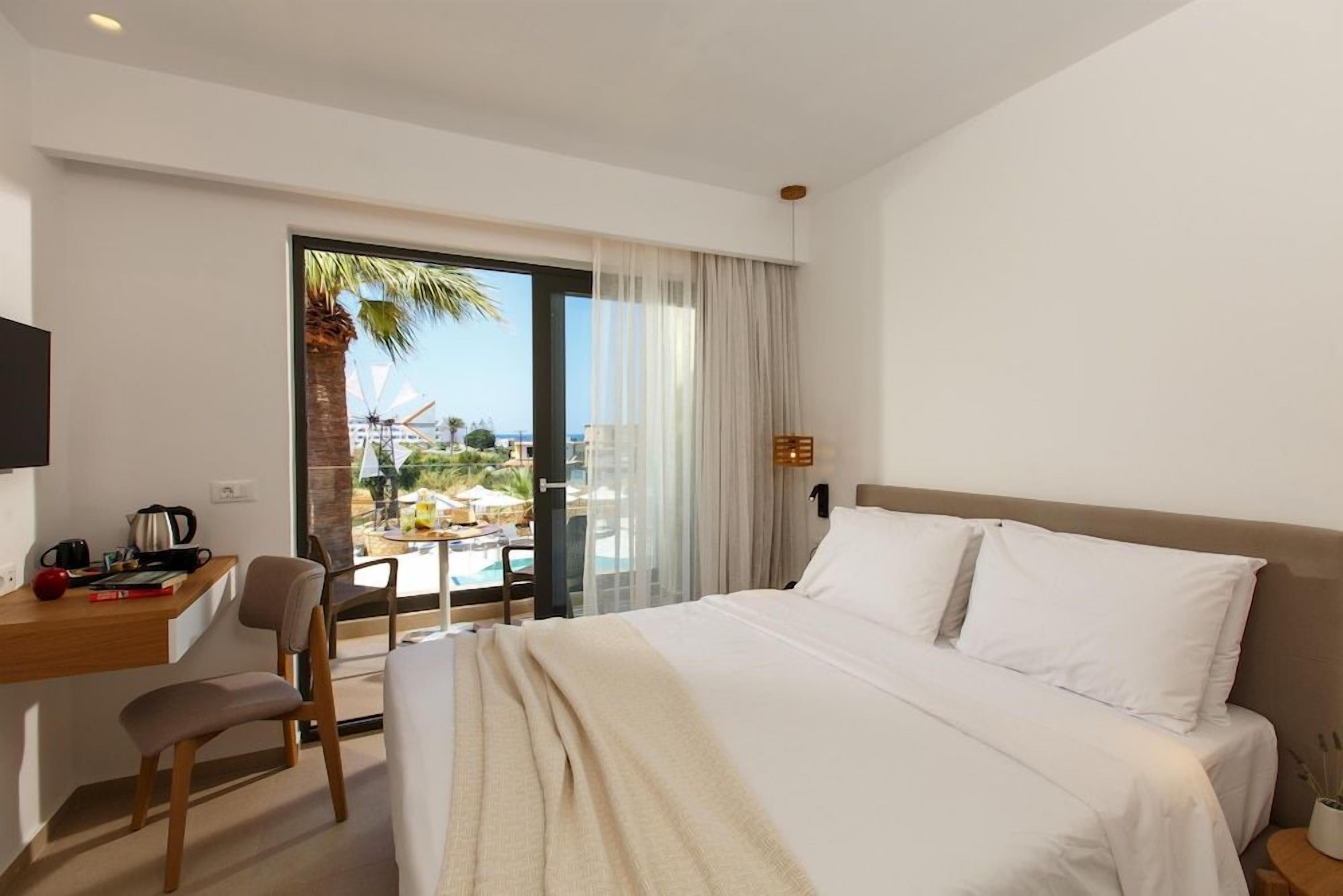 A hotel room with a bed, a desk, a chair, a TV, and a view of a pool and palm trees.